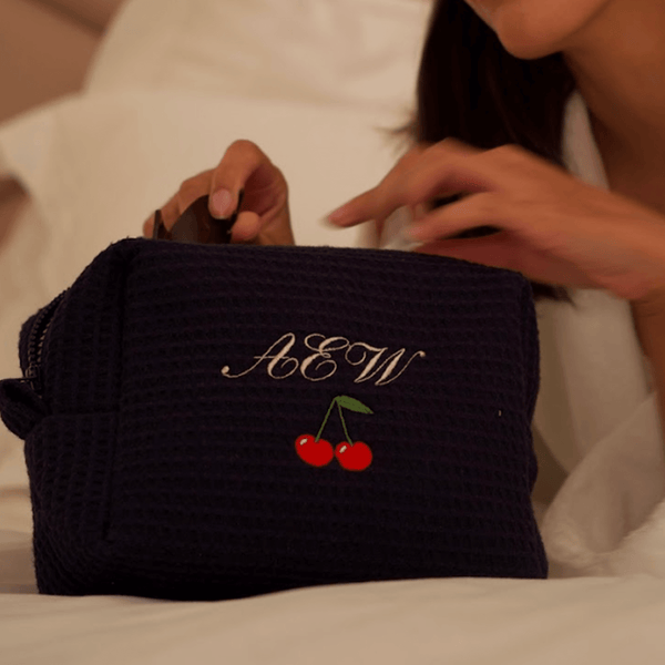 A person in a white waffle robe opens a dark waffle pouch with the initials AEW and two embroidered cherries on the front, while sitting on a bed.