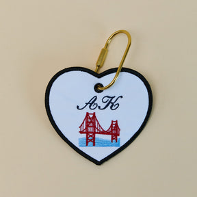 Heart-shaped keychain with a red bridge design and 'A K' initials on a beige background