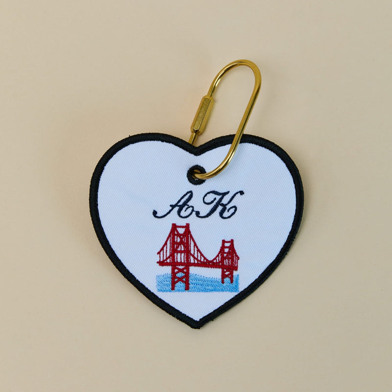 Heart-shaped keychain with a red bridge design and 'A K' initials on a beige background