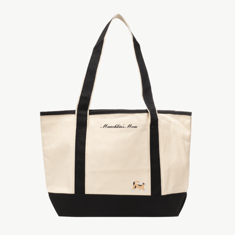 A beige canvas tote bag with long black handles and a matching black base the text 'Munchkin's Mom' near the top, and a small embroidered silhouette of a cat at the bottom right.