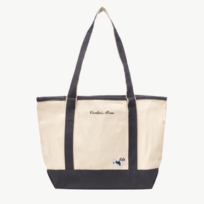 A beige canvas tote bag with long gray handles and a matching gray base the text 'Cookie's Mom' near the top, and a small embroidered silhouette of a dog at the bottom right.