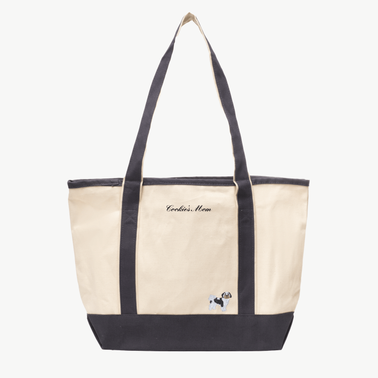 A beige canvas tote bag with long gray handles and a matching gray base the text 'Cookie's Mom' near the top, and a small embroidered silhouette of a dog at the bottom right.