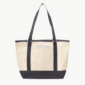 A beige canvas tote bag with gray  handles and a matching gray base. The words Surfs Up are embroidered in sky blue script on the front above the pocket. The background is plain white.