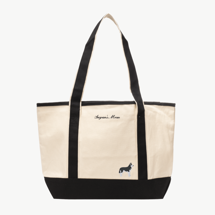 A beige canvas tote bag with long black handles and a matching black base the text 'Sagan's Mom' near the top, and a small embroidered silhouette of a dog at the bottom right.