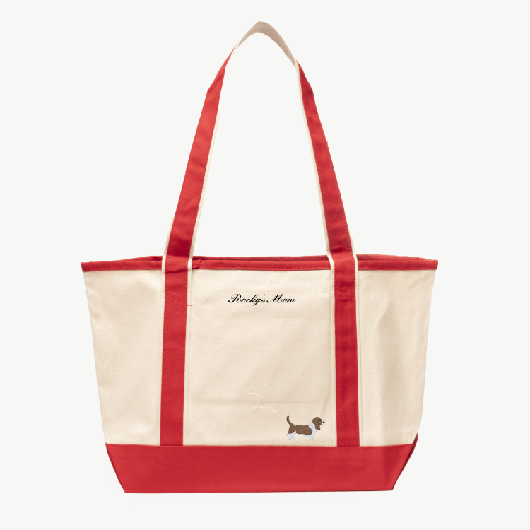 A beige canvas tote bag with long red handles and a matching red base the text 'Rocky's Mom' near the top, and a small embroidered silhouette of a dog at the bottom right.