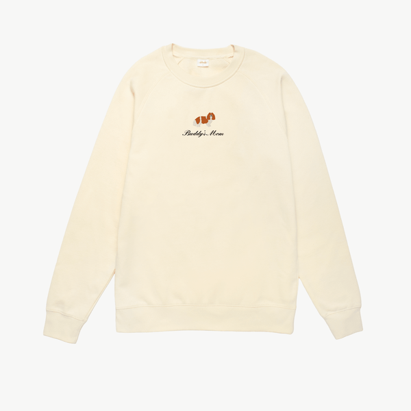 Cream crewneck sweatshirt featuring a small embroidered brown dog above the words Buddy's Mom in black script on the chest. The sweatshirt has long sleeves and a simple, relaxed design.