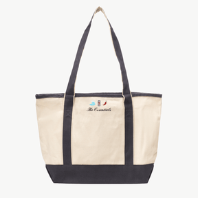 A beige canvas tote bag with long gray handles and a matching gray base, featuring three small embroidered icons on the front: a wave, a soda can, and a chili pepper. Below the icons is an embroidered text 'The Essentials'. The bag is set against a white background.