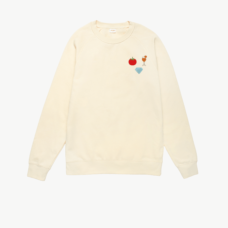 A plain cream crewneck sweatshirt with long sleeves featuring three embroidered icons of a tomato, a cocktail, and a diamond, displayed on a white background.