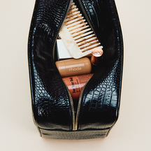 Black croc-embossed zippered pouch shown open from above, filled with hair and beauty essentials, on a cream background.