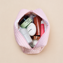 Light pink waffle zippered pouch shown open from above, filled with skincare and makeup products, on a beige background.
