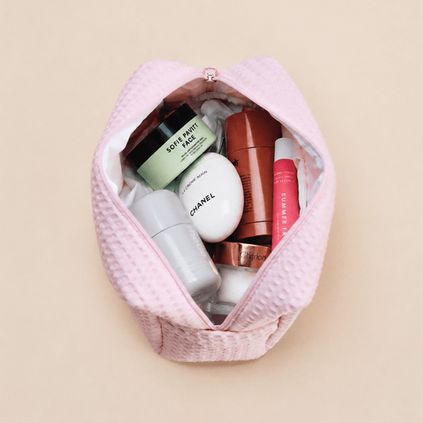 Light pink waffle zippered pouch shown open from above, filled with skincare and makeup products, on a beige background.