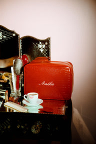 A red, croc pouch with the name Anika written in white script on the front. Set on a make up table.