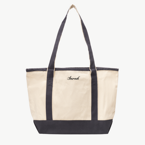 A beige canvas tote bag with long gray handles and a matching gray base. The name Sarah is embroidered in black on the front, displayed on a white background.