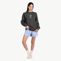 A woman stands against a white background wearing a dark gray sweatshirt with a small graphic on the chest, light blue shorts, and cream-colored socks. She has straight dark hair and a neutral expression.