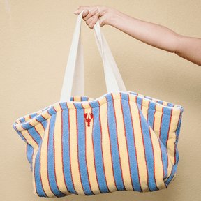 Striped beach bag with a lobster logo held by a hand against a beige background