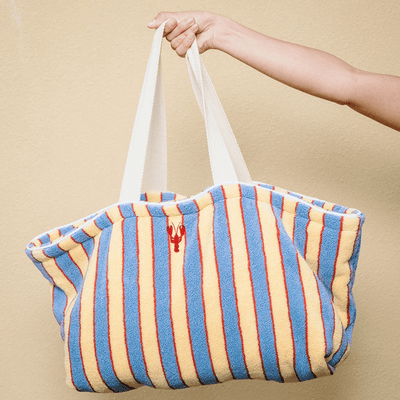 Striped beach bag with a lobster logo held by a hand against a beige background